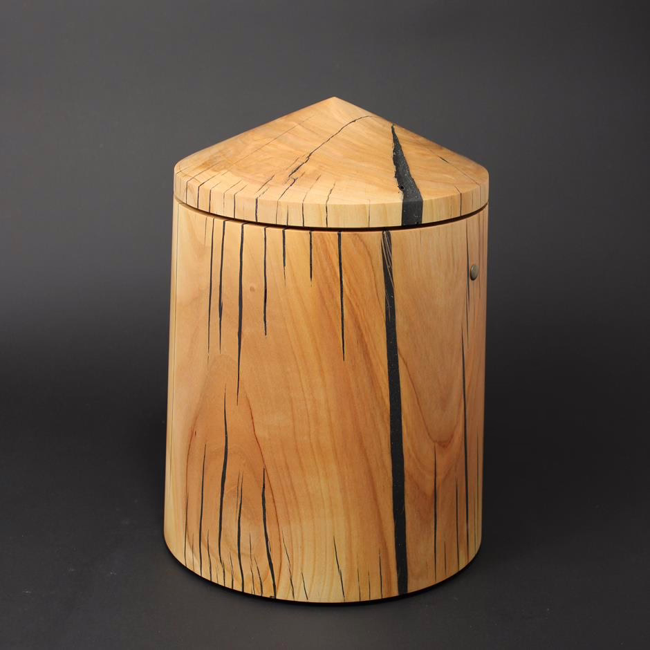 Holzurne in runder Obeliskform mit sich zur Oberseite verjüngendem Kegeldach. Holzart Kirschbaum.